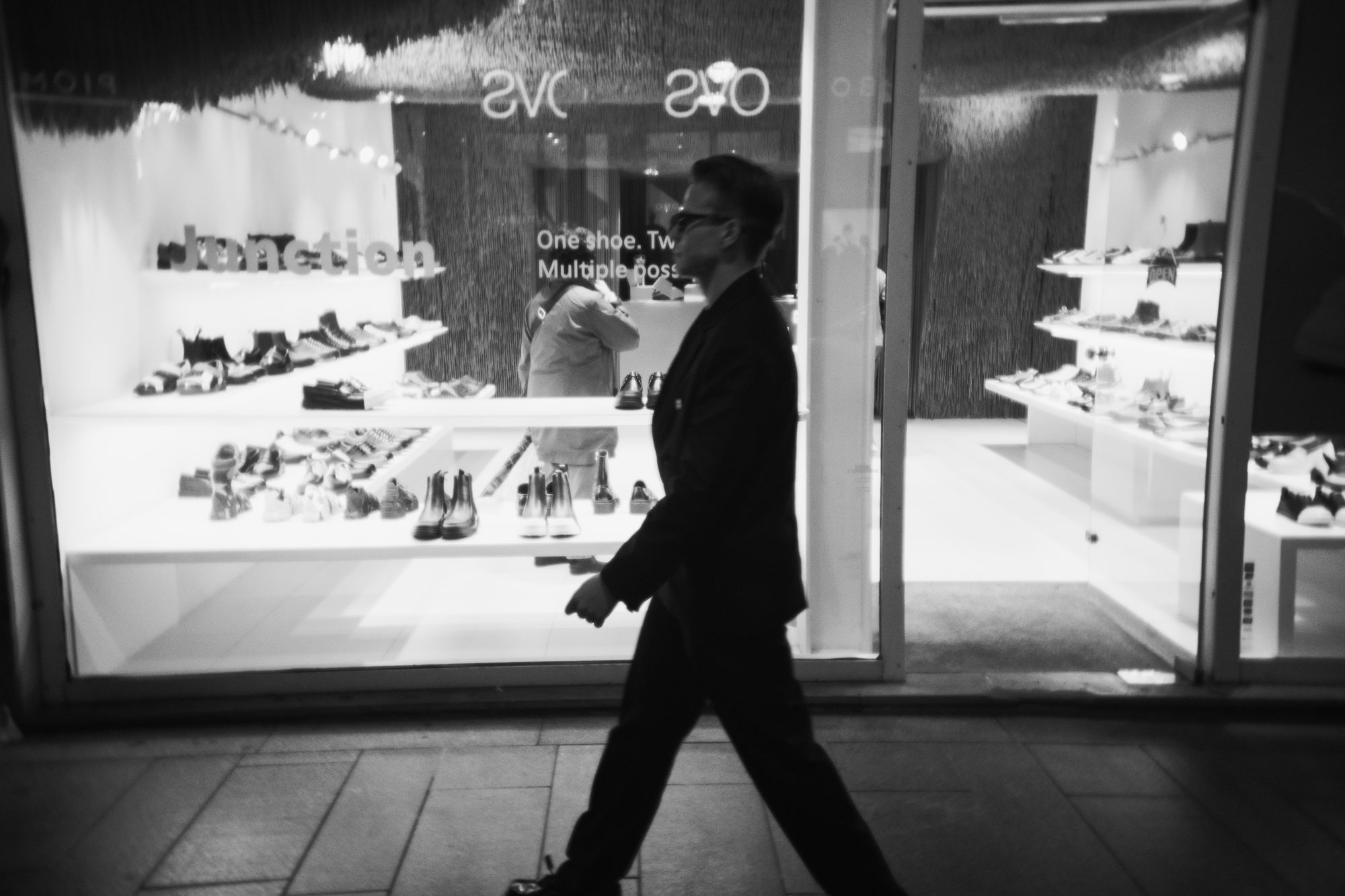 A man in a dark suit passing a lit shoe shop window at night
